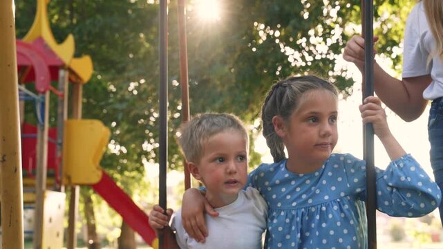 Children swing together on playground. Sibling boy and girl enjoy swinging outdoors. Kids hold swing bar in sunny park. Mother stands nearby. Happy childhood on summer playground with sunlight.