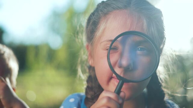 Curious girl looks through magnifying glass outdoors. Child explores nature in sunny park. Girl with braid holds lens near eye. Kid examines with curiosity and wonder. Sunlight flare glows warmly.