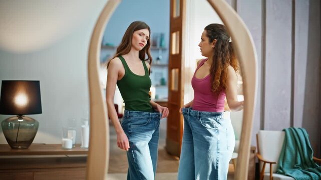 Dissatisfied friends looking mirror wearing oversize pants. Women trying jeans