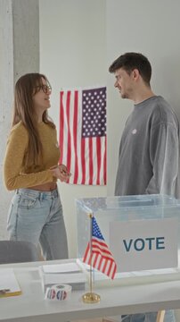 Two young voters man and woman stand by a clear ballot box with an american flag, woman clasps hands while speaking to him in a building; civic duty hopeful.