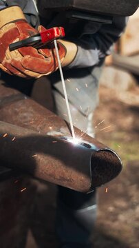 Professional worker performs arc welding on rusty metal pipe with dramatic sparks and smoke. Industrial craftsmanship and heavy manufacturing concept in warm evening light.