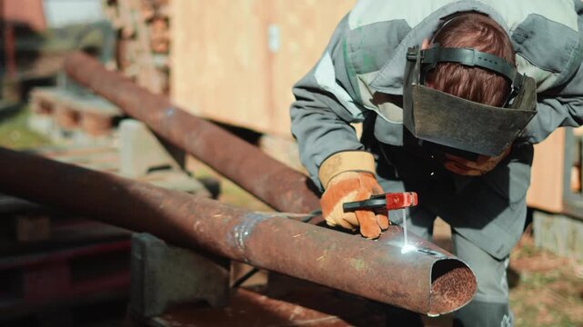 Industrial craftsman welds rusty steel pipes outside during repair process. Detailed closeup with glowing sparks and protective mask shows professional maintenance and construction expertise.