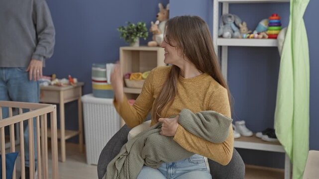 Woman cradles a swaddled baby in her lap as man leans over and gently touches her arm beside a wooden crib in a home nursery; tenderness family bonding.