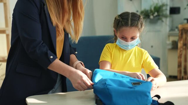 Mother helps child pack school backpack at home. Girl in face mask holds blue backpack open. Mom puts book into backpack. Parent and daughter prepare for school. Family packing school bag together.