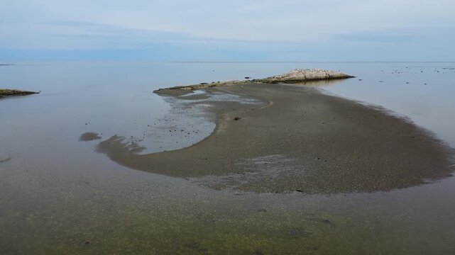 Drone footage of a minimalist seascape with curved sandbar, rocky reef, exposed mudflat and reflective low tide waters in Mitis Bay, Quebec, Canada.