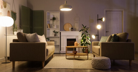 Fototapeta premium Interior of stylish living room with grey sofas, coffee table, fireplace and glowing lamps at evening