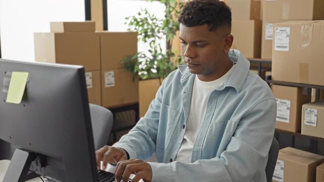 Man adjusts wrist while working at computer with parcel boxes in a building; order fulfillment focus.