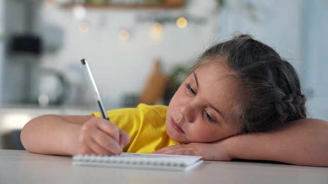 Tired child writing homework in notebook. Girl leans head on desk while writing with pen. Kid does homework in kitchen at home. Child studies and writes slowly. Little girl tired of studying at table.