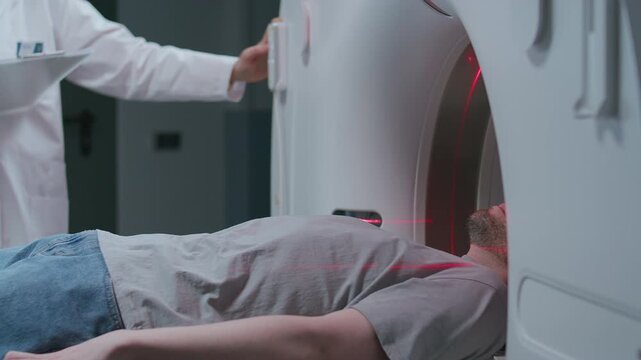 Male patient lying on motorized table as entering CT scanner, red laser lines projecting across him, technician in white medical coat operating control panel during procedure