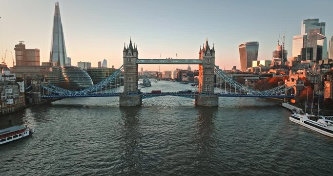 Evening urban London scene: Tower Bridge spans River Thames at golden hour, traffic cars and double decker cross the lower deck. The Shard rises among modern London skyline. Aerial view drone flight