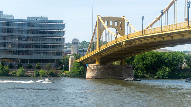 A scenic, low-angle shot of the iconic yellow Roberto Clemente Bridge (Sixth Street Bridge) stretching across the Allegheny River in downtown Pittsburgh, Pennsylvania. On the left, a modern glass-faca