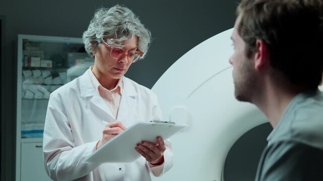 Experienced female radiologist in white lab coat taking notes on clipboard while questioning male patient before CT scan at modern diagnostic center. Over the shoulder shot