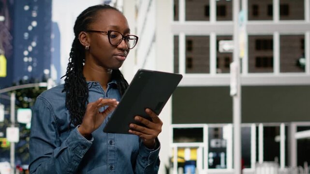 Smiling african american woman using tablet and walking in the city center, relaxing by looking at social media posts. Upbeat girl browsing navigation map in an urban landscape background.
