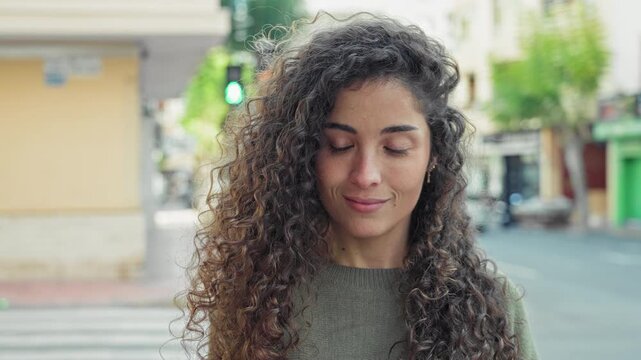 Woman face and eyes looking down with slight smile, curly hair and green sweater, standing at crosswalk street with traffic light and storefronts visible; serene contemplation.