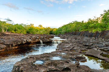 A serene waterfall cascades over rugged rocks, surrounded by lush green vegetation at sunset in Guaviare, Colombia. © Jhampier Giron
