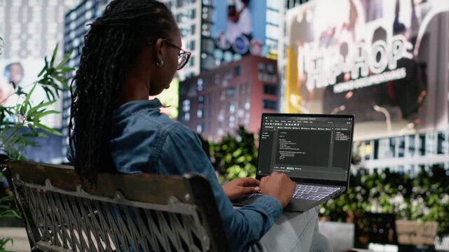 Woman software developer writing code on laptop and relaxing on park bench, implementing algorithms. Black programmer using programming language script on terminal window display.