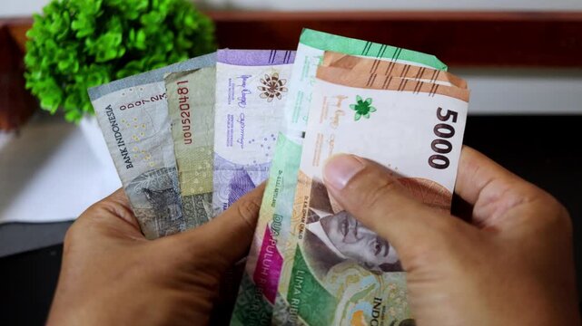 Close up of a man's hands counting Indonesian Rupiah banknotes on a wooden table.