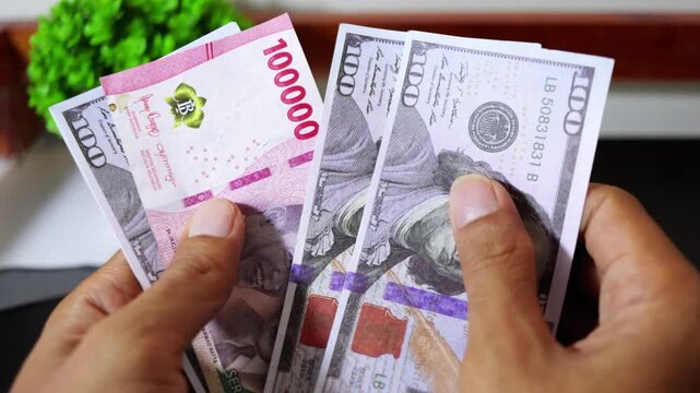 Close up of hands counting US Dollar banknotes with an Indonesian Rupiah bill among them, symbolizing currency pressure