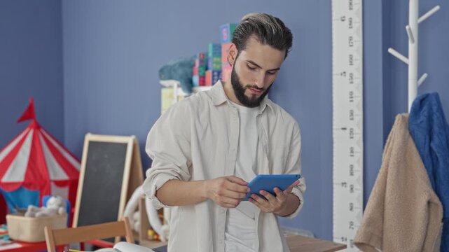 Man teacher tapping tablet with fingers in a school building surrounded by toys and learning tools; focused teaching.