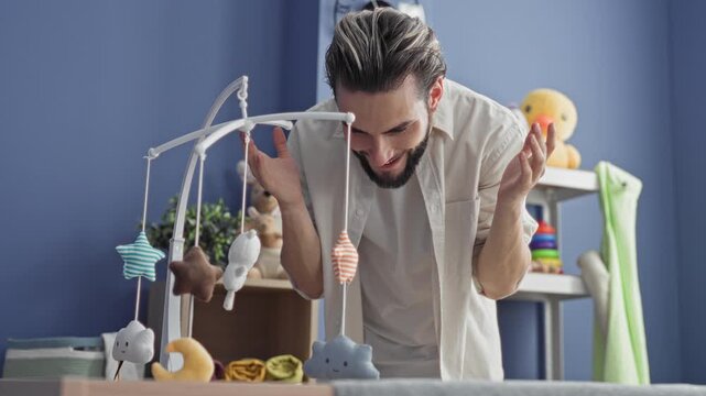 Man leans over a crib mobile, smiling with hands raised then covering his face in a nursery while playing peekaboo with the mobile; play bonding tenderness.
