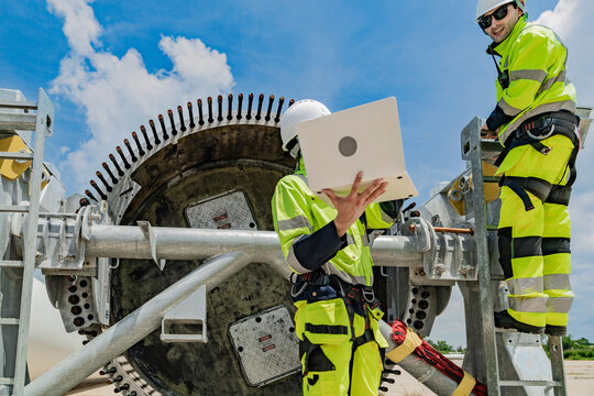 Two engineers work on a wind turbine component, with one using a laptop and the other inspecting parts on a ladder. renewable energy technology, wind power projects, and teamwork in the energy sector.