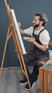 Man painting on canvas in studio, holds brush and palette with hand while seated at easel; focus creativity.