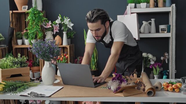 Young hispanic florist man leaning over laptop and wrapping a bouquet among potted flowers in a studio; small business focus.