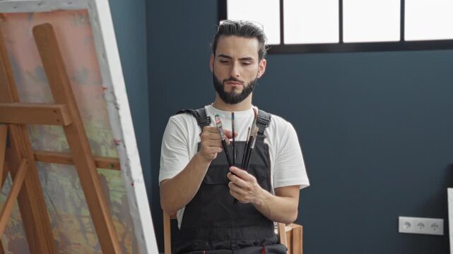 Man painter in apron inspects paintbrush tips and holds a fan of brushes near canvas in studio; focused craft.