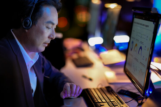 Excited corporate manager reacting to successful project results on screen. Excited man with headphones working overtime at a desk in a glowing modern workplace at night.