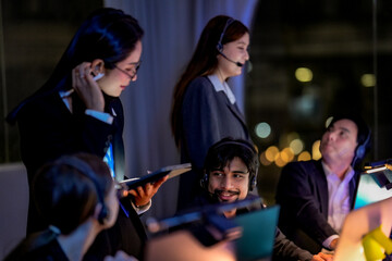 Fototapeta premium Female supervisor helping a male customer service agent during night shift. Diverse call center team working overtime in a modern dark office with beautiful city lights.