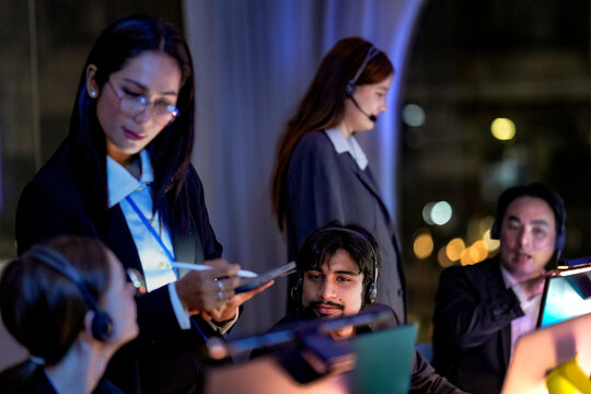 Professional businesswoman holding tablet and stylus assisting IT support agents. Multiethnic telemarketing team working overtime in a glowing workplace at night.