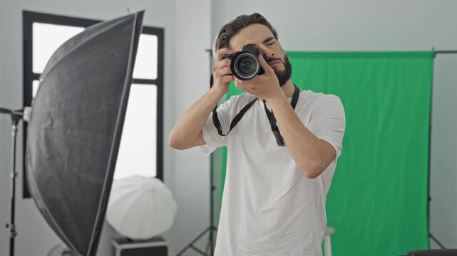 Young man photographer holds camera to eye, shows peace sign amid green screen and studio lighting with softbox and umbrella; creative playful.
