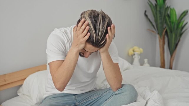 Young hispanic man seated cross legged on rumpled white bed with hands covering head and bowed gaze, plant and vase beside bed in building; anxiety fatigue introspection.