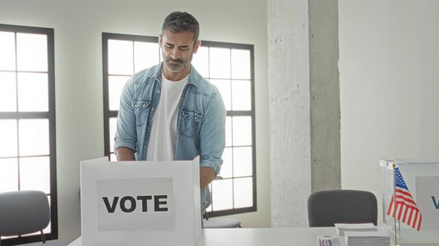 Man with hand placing ballot into voting box and holding vote button near polling table in building; civic duty pride participation.