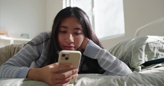 Asian Teenage girl lies on a neatly made bed, focused on a smartphone. Soft morning light from a window illuminates her striped sweater, propped on elbows face, using phone