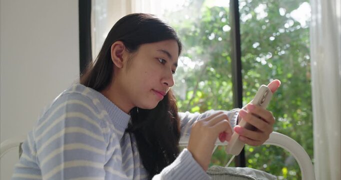 Asian teenage girl sits near a large window, using smartphone, Sunlight filters through sheer curtains in bedroom, using phone, Side view