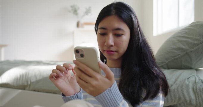 Asian teenager girl sits scrolling on her smartphone. Sunlight filters through sheer curtains onto bedroom morning, using phone