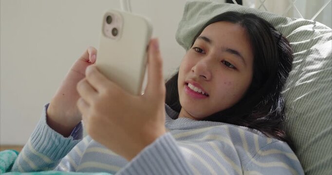 Asian teenage girl lying near on bed, using smartphone, Sunlight filters through sheer curtains in bedroom