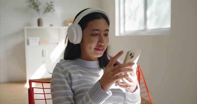 Asian teenage girl sits indoor listen to music, wearing white headphones and a light blue striped sweater, using smartphone