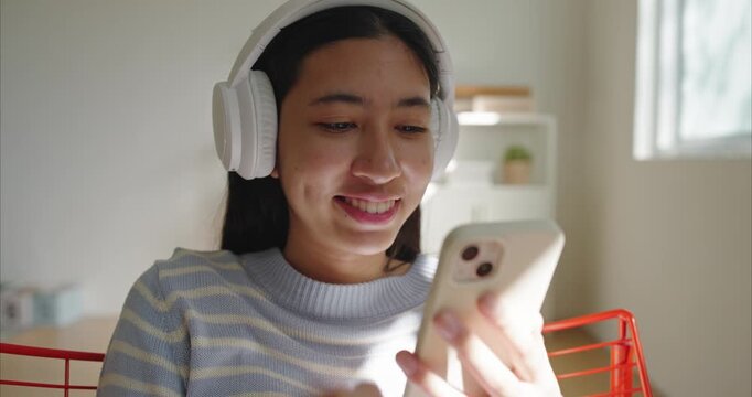 Asian teenage girl sits indoor listen to music,  wearing white headphones and a light blue striped sweater, using smartphone