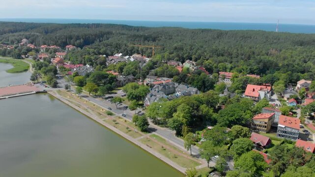 High angle, wide shot of the picturesque resort town of Nida on the Curonian Spit, Lithuania. The footage captures red roofed buildings nestled among dense green pine forests, with the calm waters of 