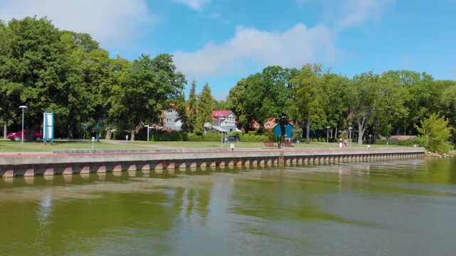 Footage capturing a beautiful, peaceful day at the seaside promenade in the resort town of Nida, located on the Curonian Spit in Lithuania. The scene features calm green water, lush trees, and buildin