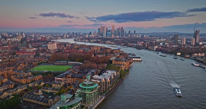 Winding River Thames flow through London urban landscape toward Canary Wharf skyscrapers under a colorful evening sky, boats and buildings in the background. Aerial view drone flight over modern city
