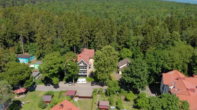 An aerial drone shot captures the charm of a small village nestled on the edge of a dense pine forest in rural Lithuania. The scene features traditional European style houses with vibrant red tiled ro
