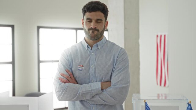 Man wearing an i voted sticker and standing with arms crossed by a transparent ballot box in a polling building with an american flag visible; determined civic duty.
