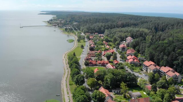 An aerial drone shot of a picturesque settlement located on the Curonian Spit, a UNESCO World Heritage site in Lithuania. The footage captures the red-roofed houses, lush green pine forests, the calm 