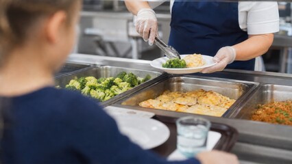  School cafeteria food service concept: Over-the-shoulder view of a child waiting in line while a worker serves a healthy hot lunch on a plate.