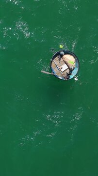 Vertical screen Aerial Hoi An Coracle Emerald Water, Lone Fisherman Rowing Circular Basket Boat, Paddle Dip, Small Net And Crates Aboard, Gentle Green Waves Shimmering Under Sunlight,