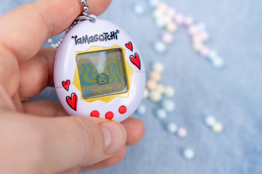 Bar, Montenegro: February 6, 2026: Hand holding white Tamagotchi virtual pet toy with pearl bead scattered on blue background, retro nostalgia youth concept.