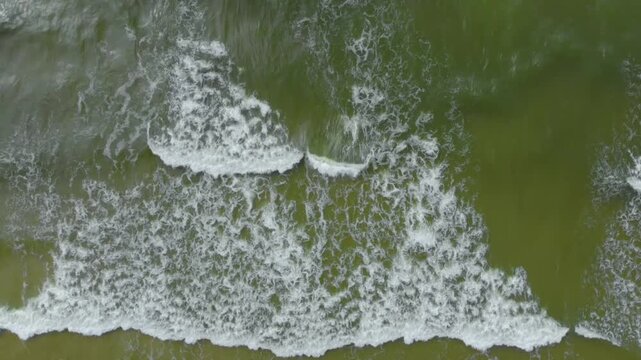 High angle, drone footage capturing the natural beauty of powerful ocean waves as they roll and break, creating white foam patterns against the greenish yellow water of the Baltic Sea.